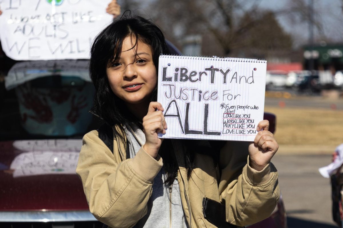 A student stands in the street holding a protest sign while other demonstrators and cars gather behind her.