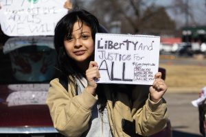 A student stands in the street holding a protest sign while other demonstrators and cars gather behind her.