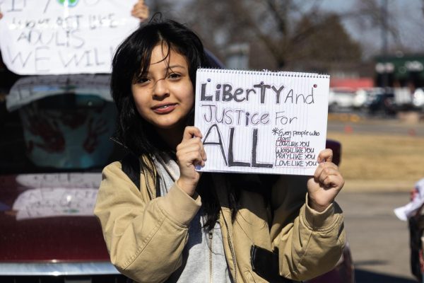 A student stands in the street holding a protest sign while other demonstrators and cars gather behind her.