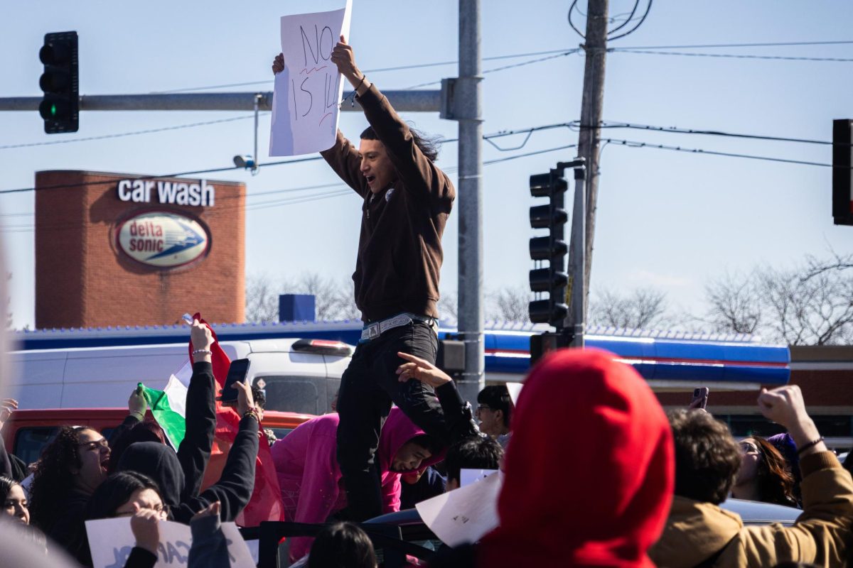 A student stands on top of a car holding a sign reading, “No One Is Illegal,” as other students climb onto the vehicle during the protest.
