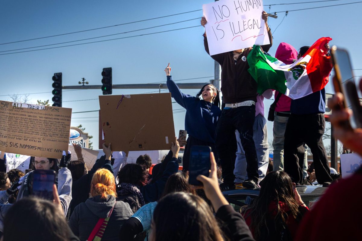 Students stand on top of a car in the middle of the street while holding signs and a Mexican flag during the anti-ICE protest.