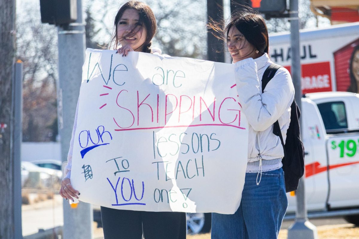 Two students stand at a street corner holding a sign that reads, “We Are Skipping Our Lessons to Teach You One!” in support of the anti-ICE walkout.